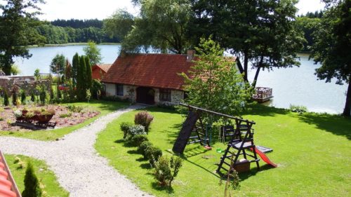 Widok na budynek kuźni, na palc zabaw i jezioro z lasem